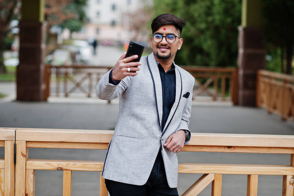Stylish indian business man with bindi on forehead and glasses, wear on gray suit posed outdoor and making selfie at mobile phone. - Фото, зображення