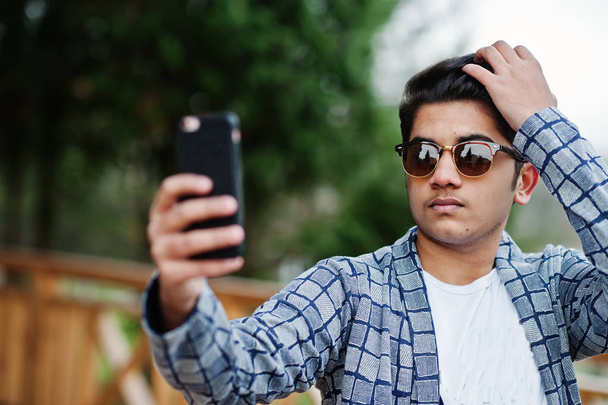 Stylish indian young man at sunglasses wear casual posed outdoor and making selfie at mobile phone. - Photo, image