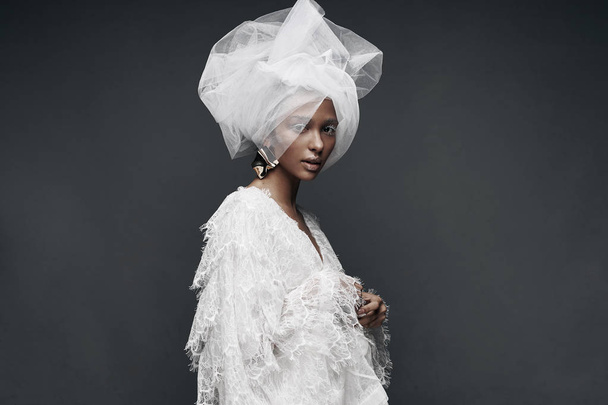 Fashion portrait of woman with white make up, in white lace dress with white veil head tie and big earring. Studio, grey background - Photo, Image