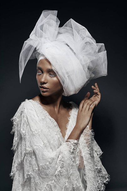Fashion portrait of woman with white make up, in white lace dress with white veil head tie and big earring. Studio, grey background - Fotó, kép