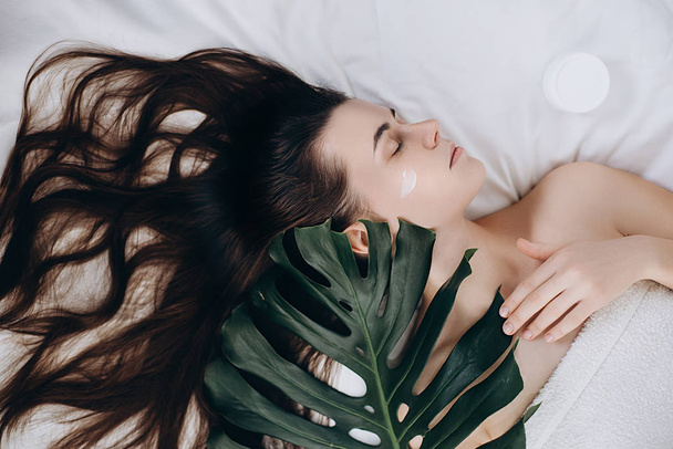 Aantrekkelijke jonge langhaar meisje crème op haar gezicht liggend op het bed met een groene blad te genieten en te ontspannen, schoonheidsinstituut toe te passen. Huid verzorging en beauty concept. - Foto, afbeelding