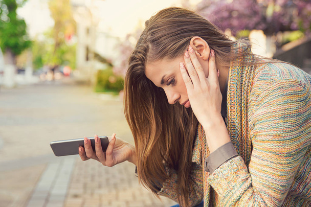 Side view of young woman holding phone and looking happy with breakup while sitting on street - Photo, image
