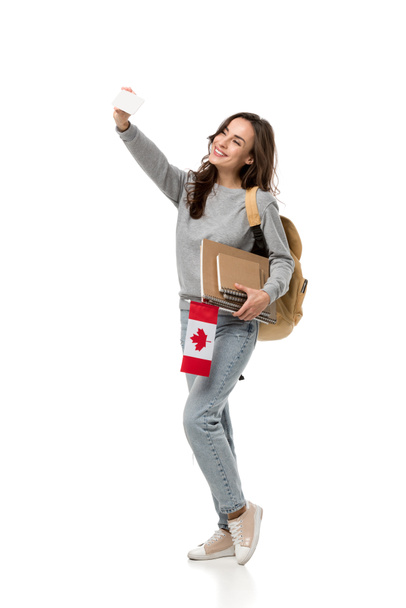 улыбающаяся студентка с блокнотами, держащими канадский флаг, делая селфи на смартфоне, изолированном на белом
 - Фото, изображение