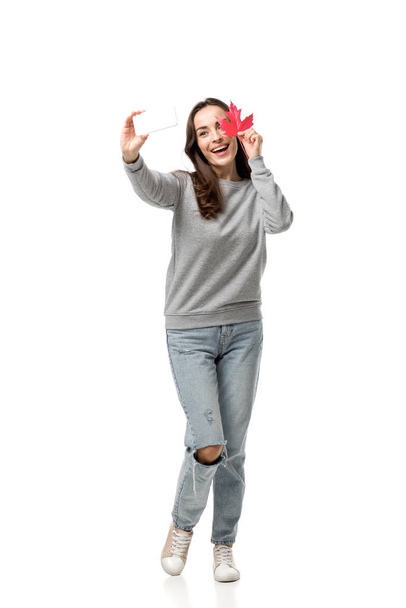カエデの葉を押しながら白で隔離のスマート フォン selfie を取ってのカジュアルな服で幸せな女 - 写真・画像