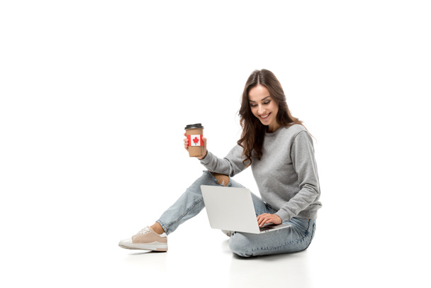 uśmiechnięta kobieta za pomocą laptopa i trzymając kubek kawy z kanadyjską flagę naklejki na białym tle - Zdjęcie, obraz