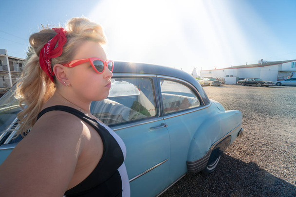 Blonde adult female with a 1950s vintage pin up hairstyle stands near an abandoned vintage car, wearing cat eye sunglasses - 写真・画像