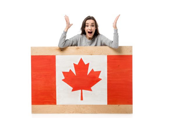 mujer sorprendida haciendo gestos con las manos detrás de una tabla de madera con bandera canadiense aislada en blanco
 - Foto, Imagen