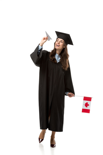 vrolijke vrouwelijke student in academische jurk holding Canadese vlag en papier vliegtuig geïsoleerd op wit - Foto, afbeelding