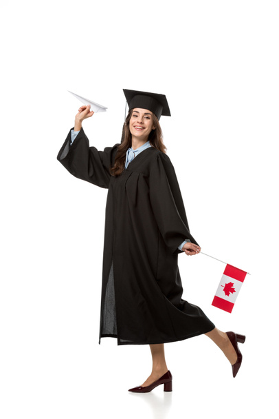 Gelukkig vrouwelijke student in academische jurk holding Canadese vlag en papier vliegtuig geïsoleerd op wit - Foto, afbeelding