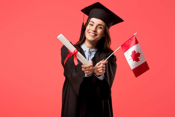 smiling female student in academic gown holding canadian flag isolated on living coral - Photo, Image
