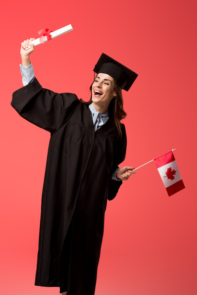 happy female student in academic gown holding canadian flag and diploma isolated on living coral - Photo, Image