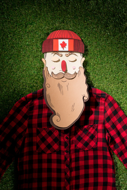 cardboard man in plaid shirt and hat with maple leaf on green grass background - Foto, afbeelding