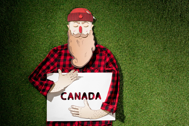 格子縞のシャツと緑の草の背景に 'カナダ' 単語とホワイト ボードを保持している帽子のダン ボール男 - 写真・画像