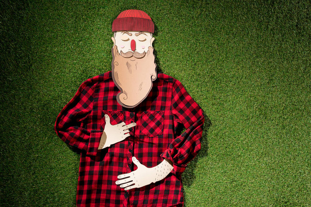 top view of cardboard man in plaid shirt and hat with maple leaf showing middle finger on green grass background - Photo, Image