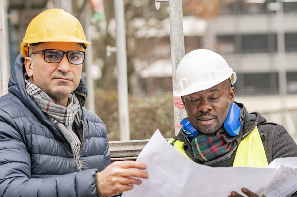 Инженеры-строители в касках и желтой куртке проверяют технические чертежи и офисные чертежи среди строительных лесов на строительной площадке
 - Фото, изображение