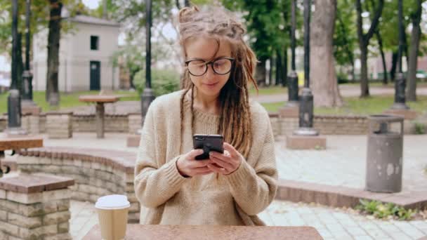 Veselá mladá Kavkazský žena s dredy má přestávka na kávu ve městě pomocí smartphone posouvání dotykové obrazovky - Záběry, video