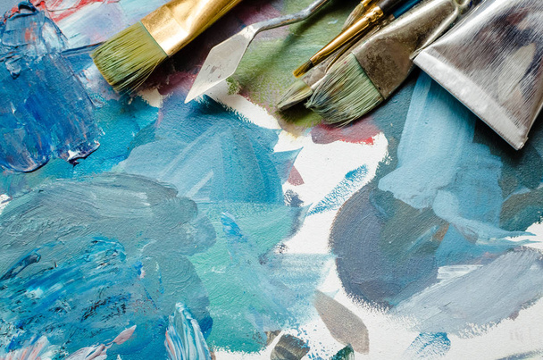 Artist paint brushes and oil paint tubes on wooden palette. Texture mixed oil paints in different colors. Instruments and tools for creative leisure. Painting hobby. Top view. Copy space. - 写真・画像