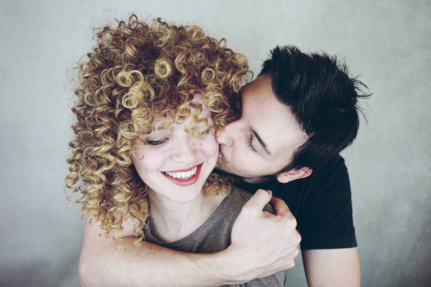 Portrait of a natural caucasian couple of young woman with curly blonde hair and man. He kisses her and she looks happy and smiles - Valokuva, kuva