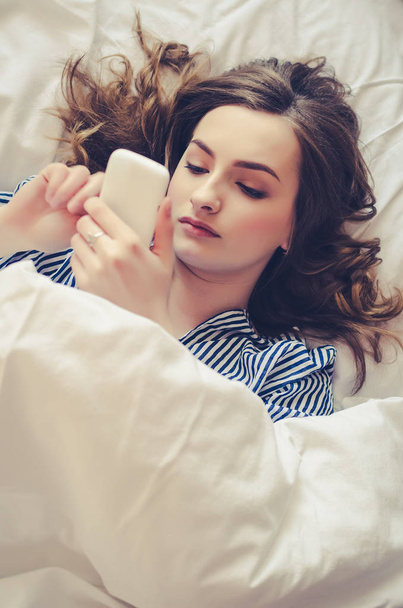 Hipster girl is reading text message from her boyfriend on mobile, while is lying in the bed. Cheerful young woman in pajamas is watching video on cell phone before of after sleeping. - Photo, Image