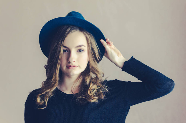 Portrait of beautiful smiling hipster teenage girl with long fair curly hair in a blue hat and black jumper looking at the camera. Lifestyle and people concept. Toned image. - Photo, Image