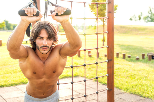 Determined young man looking serious while exercising with the suspension trainer in a modern fitness park in a sunny day of summer; trx suspension training concept; looking at camera - Photo, Image