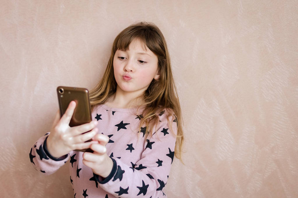 Beautiful little girl make a grimace on camera when take a selfie. Pretty child looks at the smartphone on pink background. Schoolgirl 8 years old. Copy space. - Foto, Imagem
