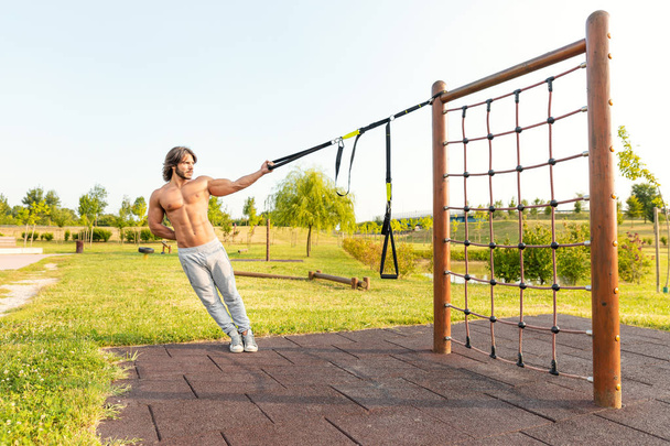 若いフィット若い男性は、健康とフィットネスの概念で側に傾いて彼の筋肉を強化し、トーンを強化するために、クライミングフレームに取り付けられた抵抗ベルトを使用して庭や公園でワークアウト。trx サスペンショントレーニングコンセプト; - 写真・画像