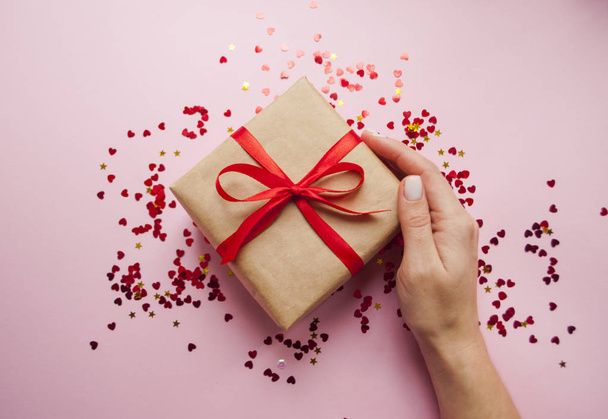 Gift box wrapped in brown colored craft paper and tied with red bow on pink background with red confetti. Lady's hand holding a giftbox. - Photo, image