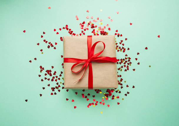 Подарункова коробка, загорнута в коричневий кольоровий ремісничий папір і зав'язана червоним бантом на бірюзовому фоні з червоним конфетті
. - Фото, зображення
