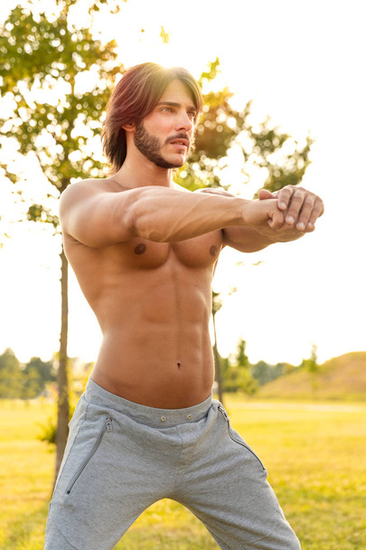 Θέα μισού μήκους ενός σέξι νεαρού άντρα με τονισμένο σώμα άσκηση τέντωμα μετά από έντονη προπόνηση στο πάρκο το καλοκαίρι - Φωτογραφία, εικόνα