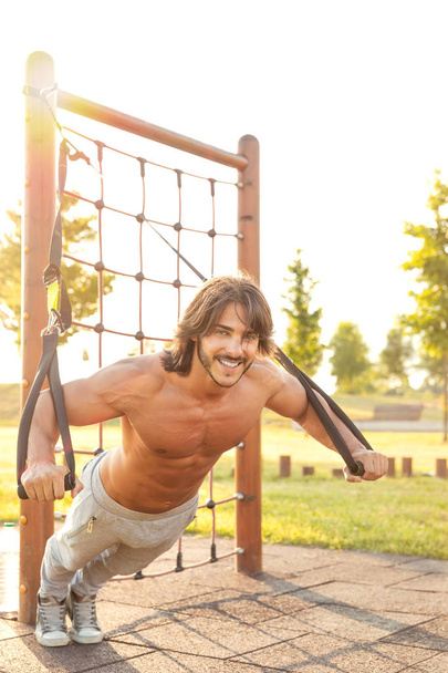 Uśmiechnięty młody człowiek szuka poważnych podczas wykonywania ekstremalnych push-up z trenerem zawieszenie w nowoczesnym parku fitness w słoneczny dzień lata; koncepcja treningu zawieszenia TRX - Zdjęcie, obraz