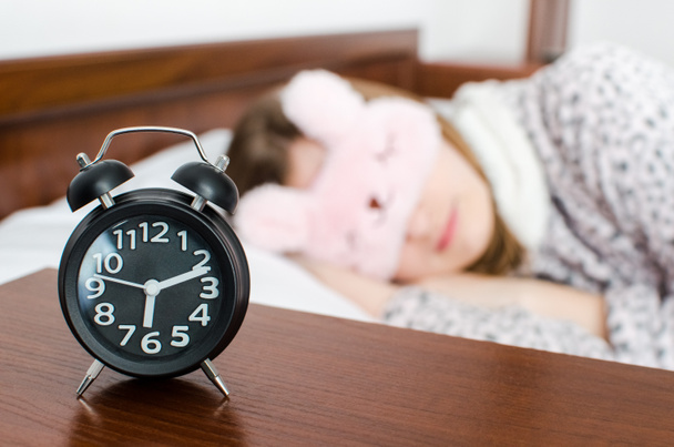 Young woman girl in cute sleep mask and warm pajamas sleep with alarm clock near the bed in the morning. Sleeping girl and alarmclock in bedroom. Teenager afraid to wake up late. Oversleep concept. - Photo, Image