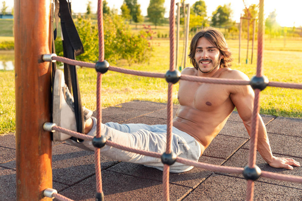 Przystojny, młody człowiek z silne ciało i brzucha sześciopak uśmiechający się podczas odpoczynku podczas treningu fitness Park w lecie; koncepcja szkoleń zawieszenie TRX - Zdjęcie, obraz