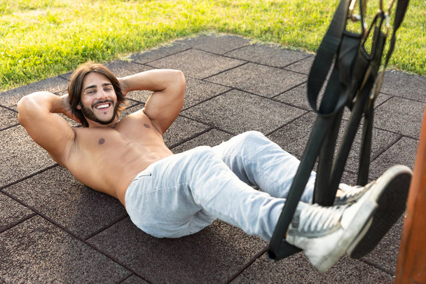 High-angle view of a smiling young man exercising crunches for the abdominal muscles during workout with the suspension trainer outdoor in the park; trx suspension training concept - Photo, Image