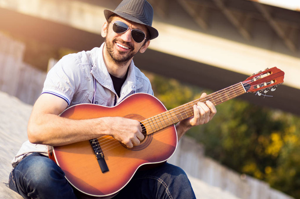 The smiling fashionable hipster guy in hat and sunglasses is playing guitar outdoors. - Fotó, kép