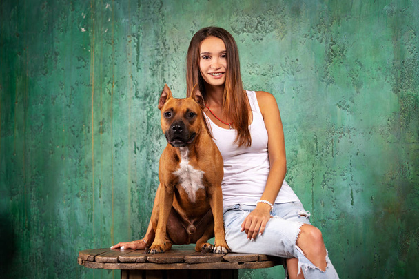Beautiful woman with her american staffordshire terrier sits indoor at green textured background - Φωτογραφία, εικόνα