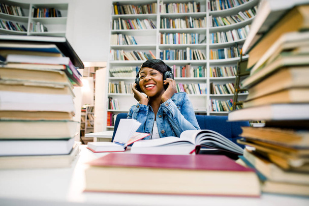 Portrait of beautiful smile black girl read books listen to earphones in library campus modern office. Young creative casual business woman working on desk with books. Successful education concept - Zdjęcie, obraz