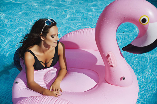 Fashionable and beautiful brunette model girl with perfect sexy body in stylish black bikini and glamorous sunglasses, posing on an inflatable pink flamingo at the swimming pool outdoors - Фото, изображение