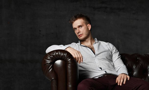 Young business man sitting in leather Chester armchair sofa in casual shirt thinking - Zdjęcie, obraz