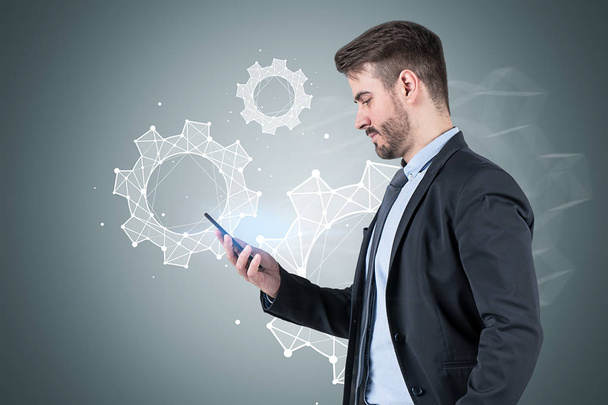 Side view of bearded young businessman looking at his smartphone standing near gray wall with cogs sketch on it. Concept of teamwork - Foto, Imagen