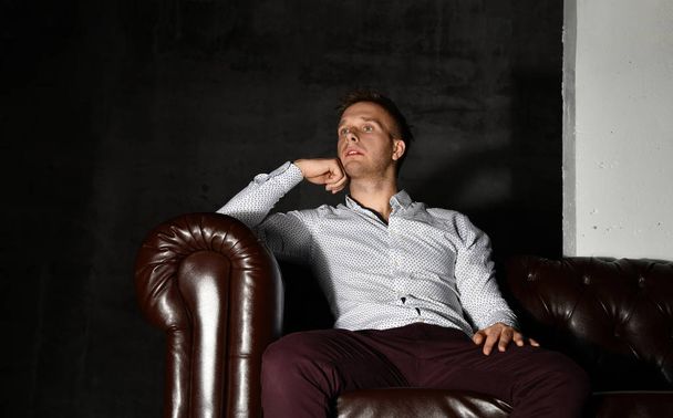 Young business man sitting in leather Chester armchair sofa in casual shirt thinking - Photo, Image
