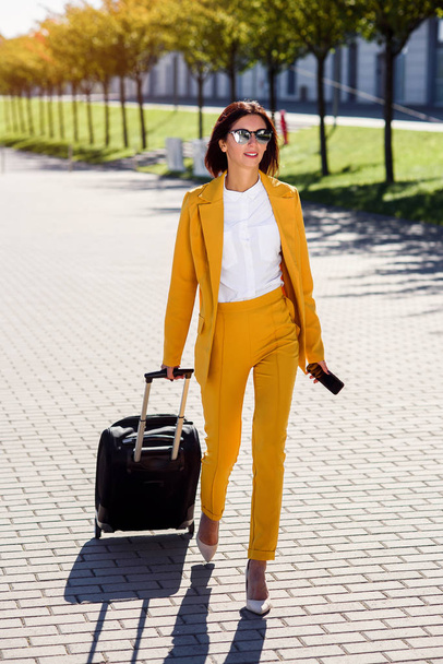 Attractive business woman in stylish yellow suit pulls a suitcase, hurries to a business meeting. Attractive business woman going on a business trip pulling her suitcase along the sidewalk behind her. - Photo, image