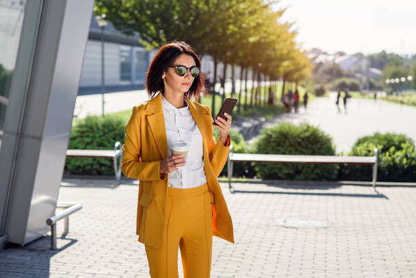 Attractive business woman in sunglasses and yellow suit walking near office centre with cup of coffee and uses smart phone. Business lifestyle and success concept. Lunch break among working day. - Photo, Image
