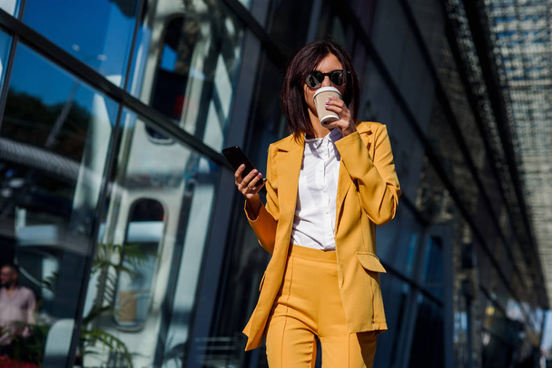 Beautiful business woman in yellow suit and sunglasses walking near business centre, drinks coffee and uses smartphone. Business concept, communication, manager and office worker. - Fotó, kép