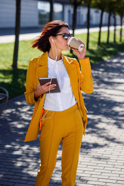 Hermosa mujer de negocios en traje amarillo y gafas de sol con tablet y auriculares bebe café mientras camina por el callejón. Mujer atractiva escucha música o audiolibro, comprueba las cartas financieras
. - Foto, imagen