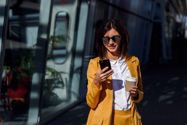 Caucasian young business woman using smart phone and drinks coffee outside near the office centre. Attractive woman scrolling pages, tapping on touchscreen, checking news and financial charts. - Zdjęcie, obraz