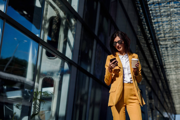 Caucasian young business woman using smart phone and drinks coffee outside near the office centre. Attractive woman scrolling pages, tapping on touchscreen, checking news and financial charts. - Fotó, kép