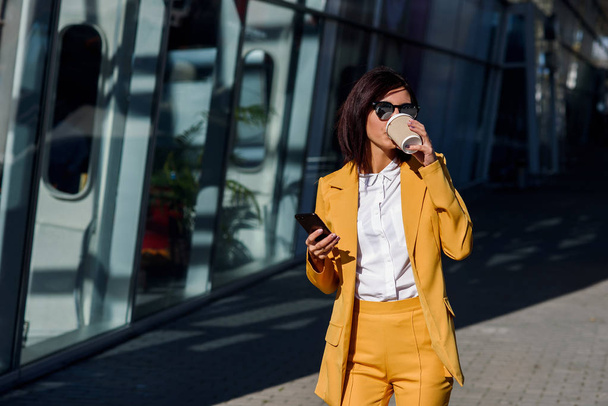 Caucasian young business woman using smart phone and drinks coffee outside near the office centre. Attractive woman scrolling pages, tapping on touchscreen, checking news and financial charts. - Foto, Imagen