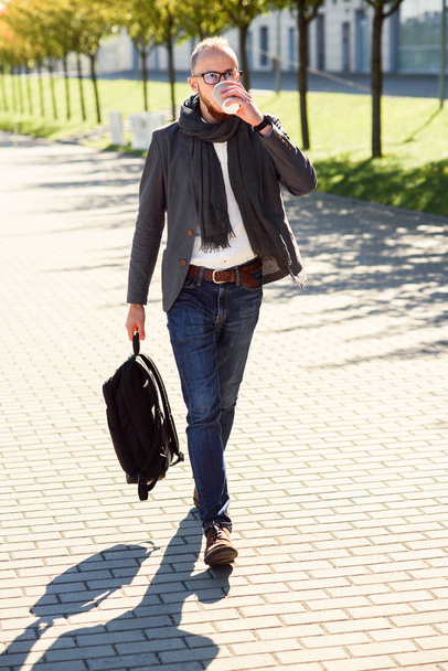 Handsome office worker in a suit with scarf holding a bag  and drinks coffee in the morning before work start near office building. Young businessman going to business meeting. - Foto, immagini