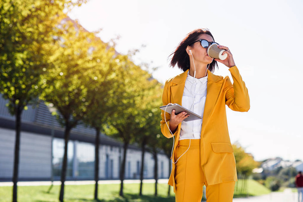 Stylish business woman in yellow suit with tablet computer and headphones drinks coffee while walks on the alley. Attractive woman listens music or business news, checks financial charts. - Фото, изображение